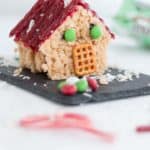 edible 3D house made of christmas rice krispie treats, red licorice roof shingles, mint M&M windows, pretzel doorway, and coconut snowflakes.
